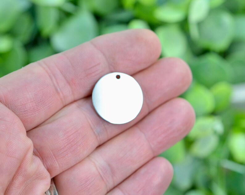 May include: A small, circular, silver-colored metal disc charm with a smooth, reflective surface. The disc features a single, centered circular hole near the top edge, designed for attachment to jewelry or other decorative items.