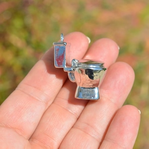 May include: A silver-colored toilet charm pendant. The charm is shaped like a toilet with a closed lid. The charm is being held in a hand.