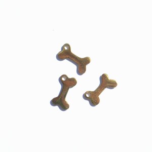 May include: Three small, gold-colored bone charms are scattered on a white surface. Each charm has a bone shape with a small hole at one end. The charms are casting shadows, indicating a bright light source.