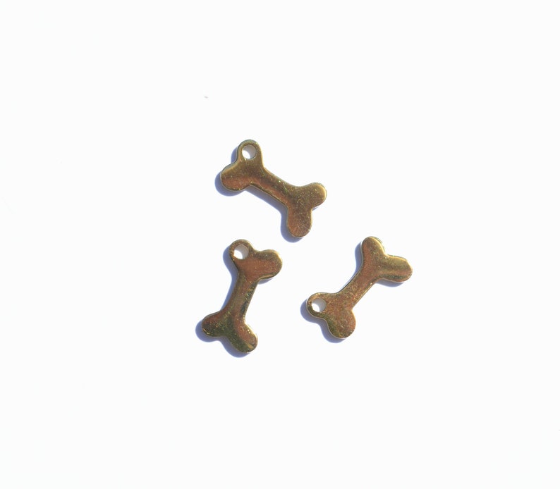May include: Three small, gold-colored bone charms are scattered on a white surface. Each charm has a bone shape with a small hole at one end. The charms are casting shadows, indicating a bright light source.