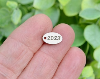 May include: A small, oval-shaped silver-toned metal charm featuring the engraved number 2023 and a single circular hole on the left side for attachment. The charm has a smooth, polished surface and is presented in a clear, focused view.
