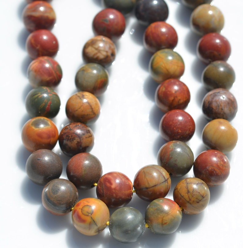 Puede incluir: Una hebra de cuentas redondas de piedras preciosas multicolores. Las cuentas son una mezcla de marr&oacute;n, rojo y amarillo con patrones en espiral. Las cuentas est&aacute;n enhebradas en un hilo dorado.