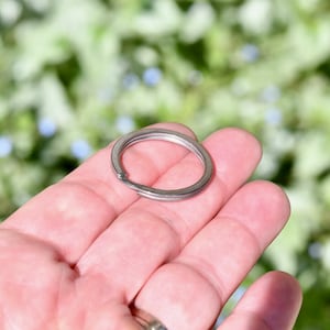 May include: A silver key ring with a circular shape, held in a hand against a blurred green background.