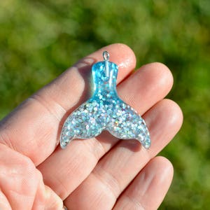 May include: A light blue mermaid tail charm with silver glitter embedded in the resin. The tail is a pendant and has a silver loop at the top for attaching to a necklace or bracelet.