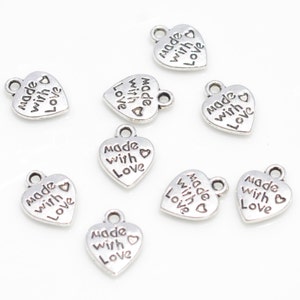 May include: A collection of small, silver-colored heart-shaped charms. Each charm has the words "made with love" engraved on it, along with a small heart symbol. The charms have a loop at the top for attaching to jewelry or other crafts.