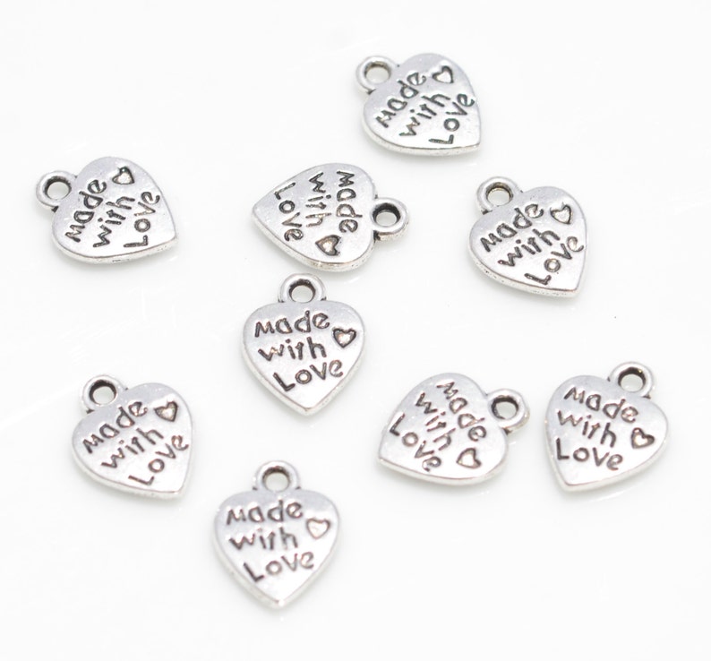 May include: A collection of small, silver-colored heart-shaped charms. Each charm has the words "made with love" engraved on it, along with a small heart symbol. The charms have a loop at the top for attaching to jewelry or other crafts.