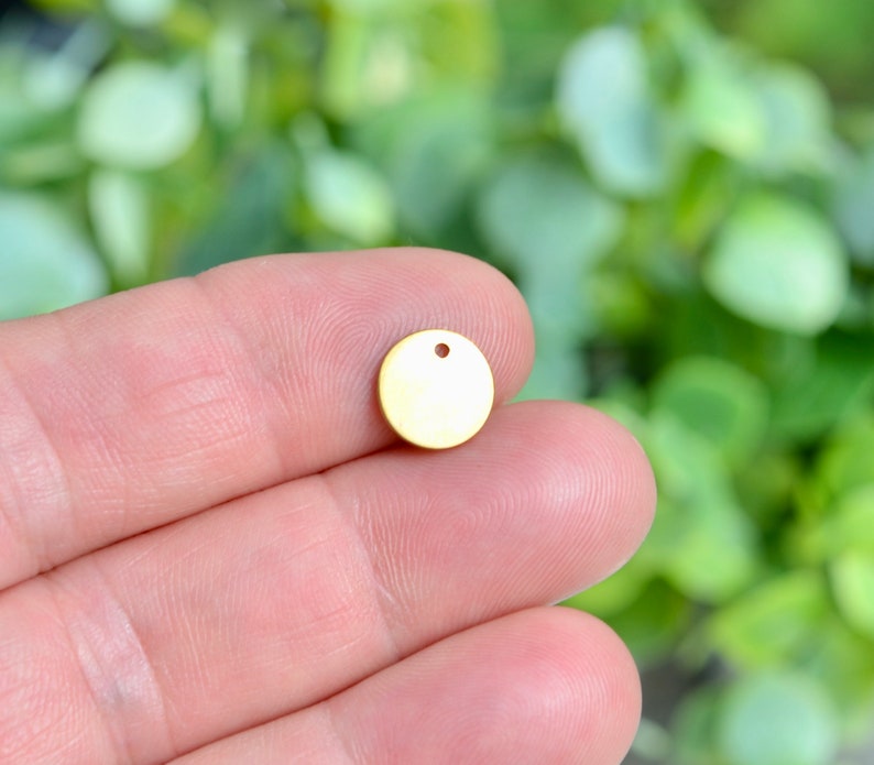 Op de afbeelding: Een gouden metalen cirkel met een klein gaatje in het midden. De cirkel heeft een diameter van ongeveer 2,5 cm.