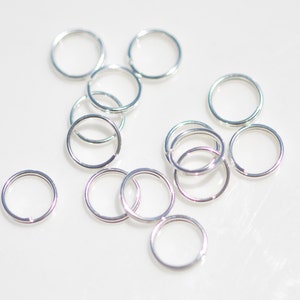 May include: A collection of silver jump rings, a type of jewelry finding, scattered on a white surface.