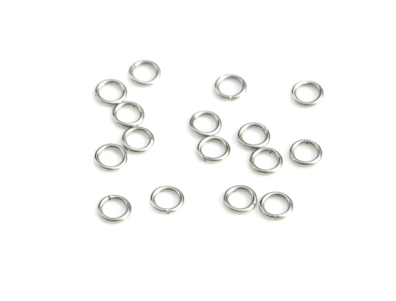 May include: A collection of silver jump rings, scattered on a white background. The rings are small and circular, with a smooth, metallic finish.