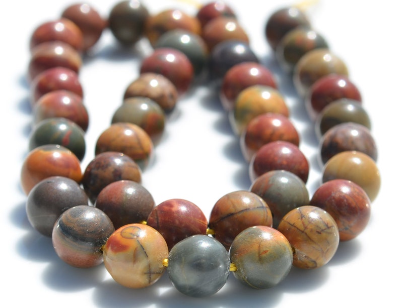 Puede incluir: Una hebra de cuentas de piedras preciosas multicolores. Las cuentas son redondas y tienen un aspecto moteado con tonos de marr&oacute;n, rojo y gris. Las cuentas est&aacute;n enhebradas en un cord&oacute;n transparente.