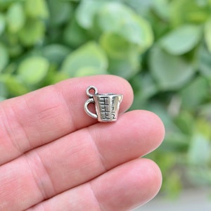 May include: Silver measuring cup charm with a handle and markings for 1/2, 1, 1 1/2, and 2. The charm is on a finger.