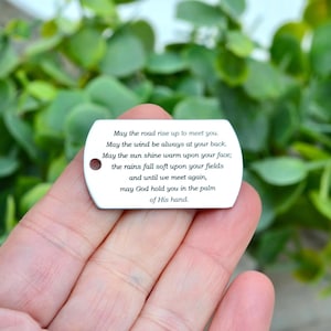 May include: A small, silver tag with rounded edges and a hole at the top. The tag has engraved text of a blessing. The background is a blurred view of green foliage.