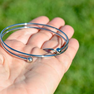 May include: A silver bangle bracelet with two round beads. The bracelet is open and has a simple design.