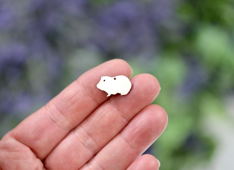 May include: White metal charm shaped like a guinea pig. The charm is small and has a hole for attaching to a necklace or bracelet.