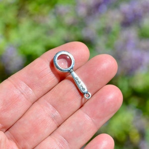 May include: A silver magnifying glass charm with a decorative handle. The charm is small and would be perfect for adding to a necklace or bracelet.