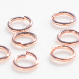 May include: Ten rose gold jump rings, a type of jewelry finding, are arranged on a white surface. The rings are all the same size and are made of a smooth, shiny metal.
