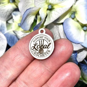 May include: A small, circular, silver charm with the words "SUPPORT YOUR LOCAL CROCHETER" around a central design of crochet hooks and yarn. The charm has a small hole at the top. The background is blurred, with blue and white flowers.