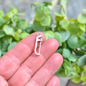 May include: A small, silver-colored saw charm with a loop at the top for attaching to jewelry or other crafts. The saw blade has detailed teeth, and the handle is a simple, curved design. The charm is held in a hand against a blurred green background.