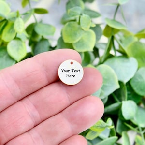 May include: A small, round, cream-colored charm with a hole at the top. The charm has the words "Your Text Here" in black, centered on the surface. The charm is held in a hand, with a background of green foliage.