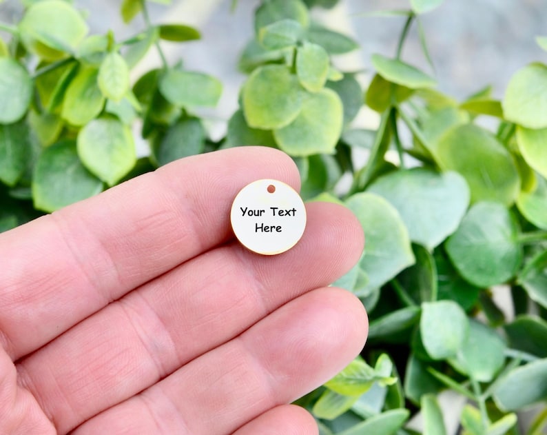 May include: A small, round, cream-colored charm with a hole at the top. The charm has the words "Your Text Here" in black, centered on the surface. The charm is held in a hand, with a background of green foliage.