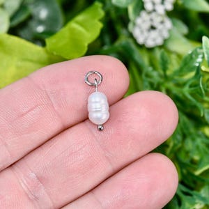 May include: A small, white, oval pearl charm with a silver loop and a small silver bead. The pearl has a ridged texture. The charm is held in a person's hand against a backdrop of green foliage.