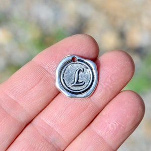 May include: Silver charm with a raised letter L in a circular design. The charm has a hole at the top for attaching to a necklace or bracelet.