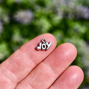 May include: A small, silver-colored charm spelling out the word "JOY". The charm is in a simple, block letter style with a small loop at the top for attaching to a bracelet or necklace. The charm is held between fingers against a blurred green background.
