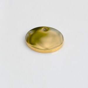 May include: A gold-colored, round pendant with a slightly domed surface and a small hole at the top. The pendant has a polished finish and a raised edge. The object is set against a white background.