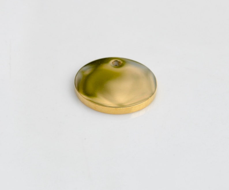 May include: A gold-colored, round pendant with a slightly domed surface and a small hole at the top. The pendant has a polished finish and a raised edge. The object is set against a white background.