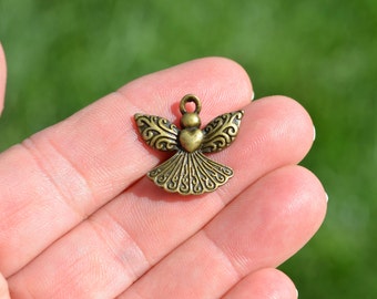 May include: A bronze angel charm with a heart-shaped body and intricate details. The wings are spread out and the charm has a loop at the top for attaching to a necklace or bracelet.