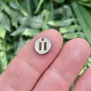 May include: A silver charm with a round shape and a raised design of two vertical lines. The charm has a small hole at the top for attaching to a bracelet or necklace.