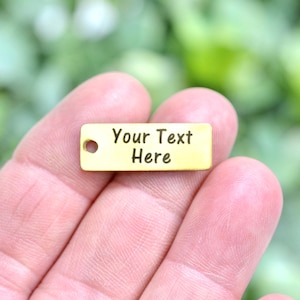 May include: A small, rectangular gold-coloured charm with the text "Your Text Here" engraved in black. The charm has a small hole on the left side. The background is a blurred green.