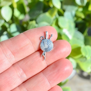 May include: Silver charm featuring a ball of yarn with two knitting needles. The charm is a popular choice for jewelry making and crafting projects.