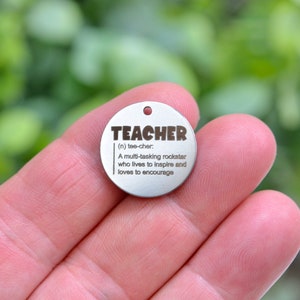 May include: A silver round charm with the word "TEACHER" and a definition: "(n) tee-cher: A multi-tasking rockstar who lives to inspire and loves to encourage."