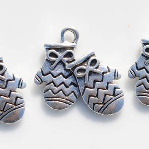 May include: Three silver-toned mitten charms with a bow detail and a zig-zag pattern. Each charm has a loop at the top for attaching to a necklace or bracelet. The mittens are a winter holiday theme.