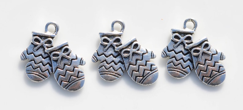 May include: Three silver-toned mitten charms with a bow detail and a zig-zag pattern. Each charm has a loop at the top for attaching to a necklace or bracelet. The mittens are a winter holiday theme.
