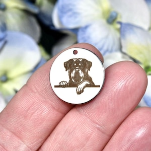 May include: A circular silver-toned metal pendant featuring a dark brown laser-engraved illustration of a Rottweiler dog resting its paws on a horizontal line. The pendant has a small hole at the top for attachment to a chain or cord.