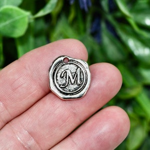May include: Silver charm with a wax seal design featuring the letter "M" in a raised, stylized font.