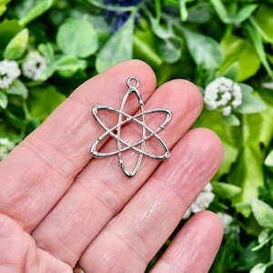 May include: A silver atom charm with a loop for attaching to jewellery. The charm is held in a hand against a backdrop of green and white foliage. The charm is a science-themed jewellery component, suitable for necklaces or bracelets.