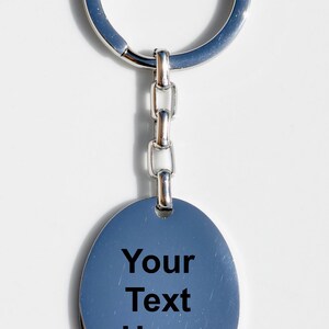 Puede incluir: Llavero plateado con un colgante ovalado. El colgante tiene el texto "Your Text Here" en negro. El llavero incluye un anillo y un eslab&oacute;n de cadena. El fondo es blanco.