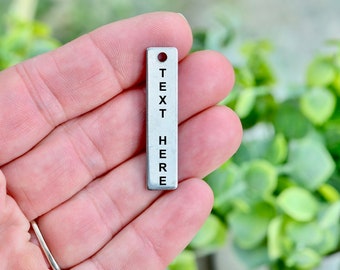 May include: A small, rectangular silver charm with a hole at the top. The charm has the words "TEXT HERE" vertically stamped on it in black, centered on the surface. The background is a soft focus of green foliage.