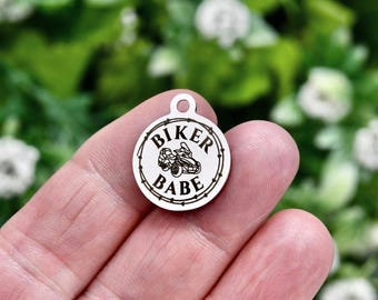 May include: A circular silver-toned metal charm featuring a small loop at the top. The center displays an engraved graphic of a three-wheeled motorcycle surrounded by a circular border, with the text BIKER BABE printed in black capital letters.
