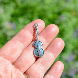 May include: Silver charm in the shape of a violin. The charm is small and detailed, with a classic violin design.