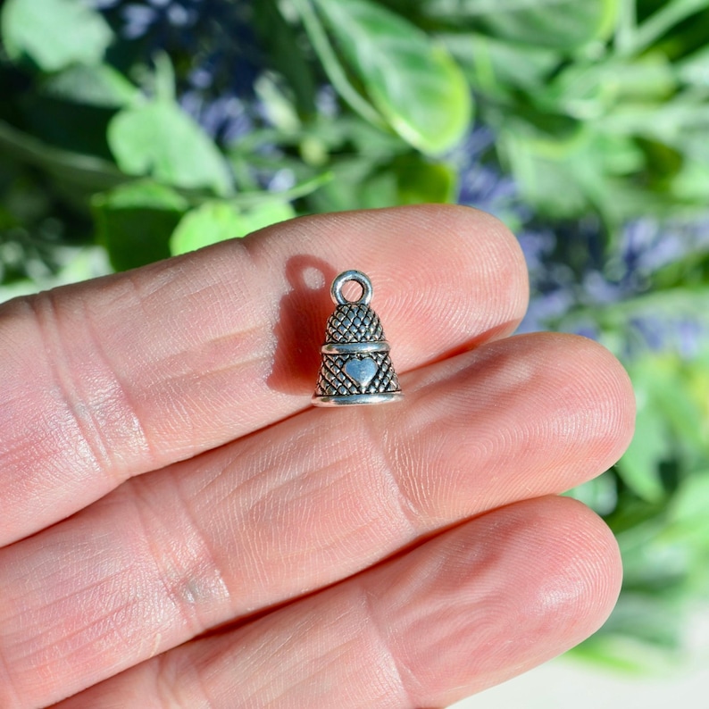 Peut inclure: Une petite breloque en m&eacute;tal argent&eacute; en forme de d&eacute; &agrave; coudre, pr&eacute;sentant un motif textur&eacute; en croisillons sur toute sa surface et un c&oelig;ur lisse en relief centr&eacute; sur le devant. La breloque comporte une petite boucle circulaire au sommet.
