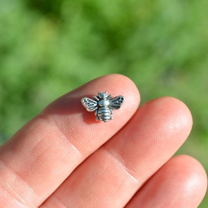 May include: A small, detailed silver-colored bee charm with outstretched wings. The bee is resting on a finger, with a blurred green background. This charm could be used for jewelry making or other crafts.