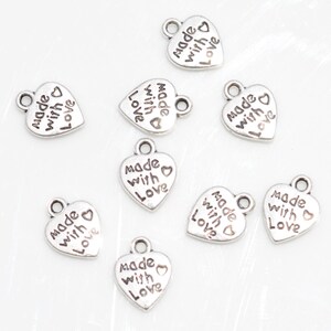 May include: A collection of small, silver-colored heart-shaped charms. Each charm has the words "made with love" inscribed on it. The charms have a loop at the top for attaching to jewelry or other crafts.