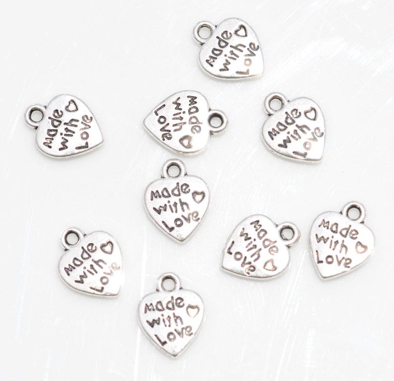 May include: A collection of small, silver-colored heart-shaped charms. Each charm has the words "made with love" inscribed on it. The charms have a loop at the top for attaching to jewelry or other crafts.