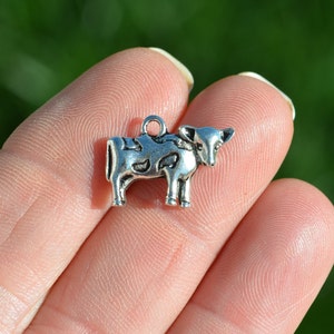 May include: Silver charm in the shape of a cow with black spots. The charm is on a finger.