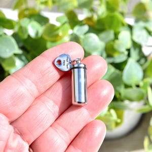 May include: A small, silver-colored cylindrical pendant with a heart-shaped tag. The tag has a small hole and the words "Your Text Here". The pendant is held in front of a background of green foliage.