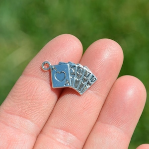 May include: Silver charm of a hand of playing cards, including the Ace of Hearts, King, Queen, Jack, and 10. The charm is on a finger.